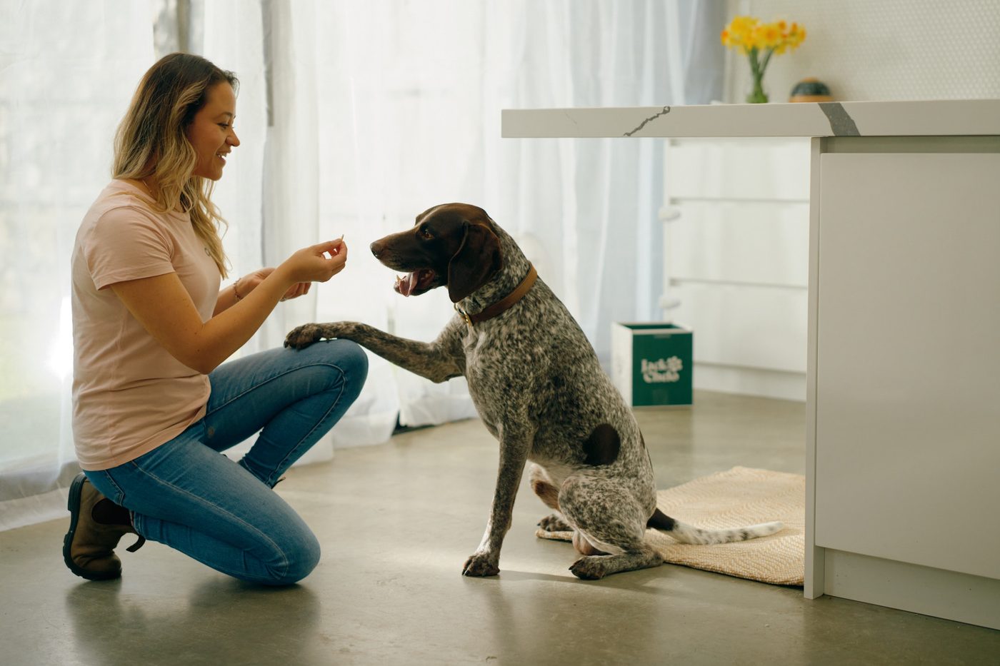 A woman kneeling on the kitchen floor as her dog offers a paw, with a Jack and Chelo box nearby