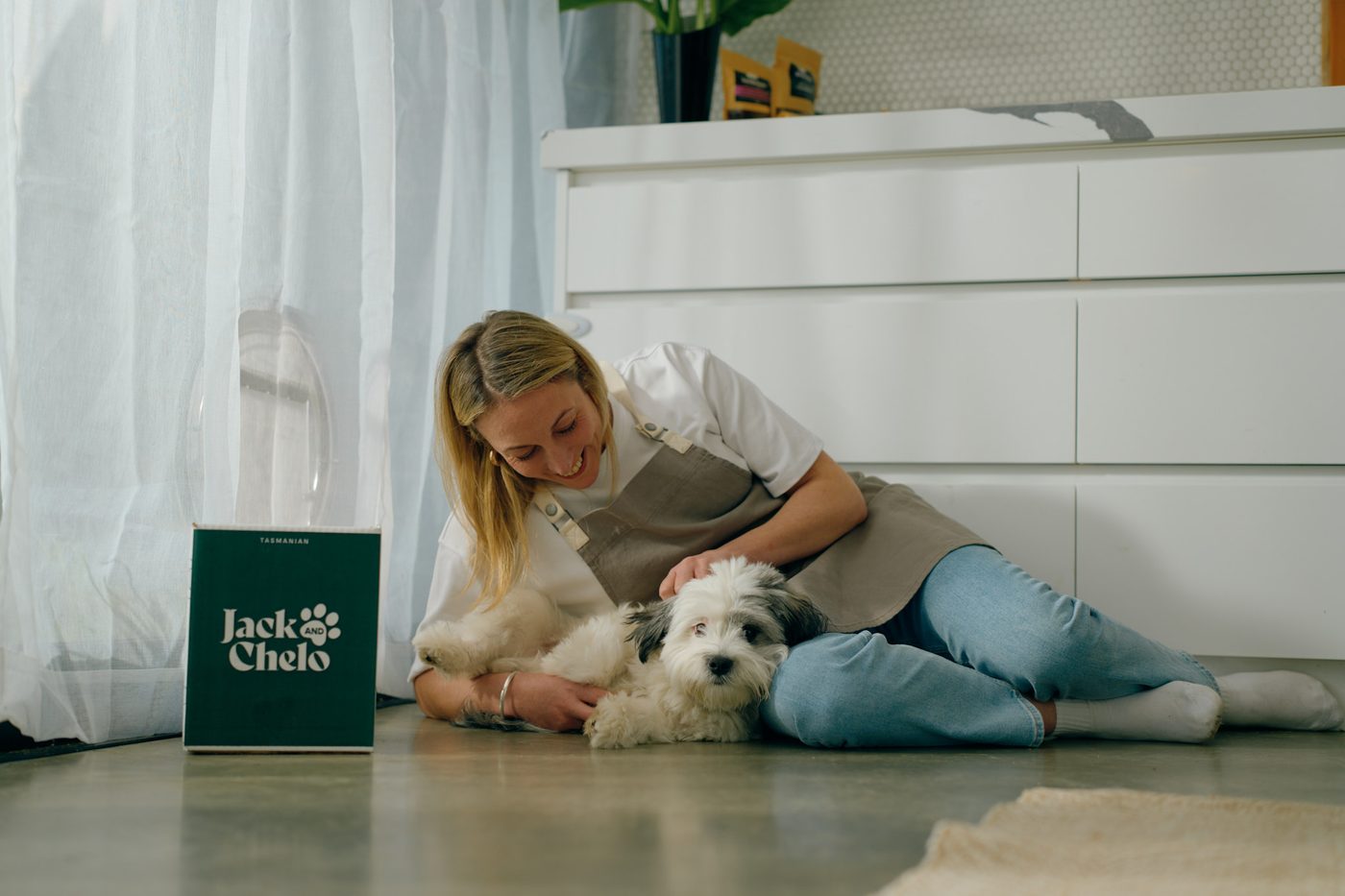 A woman lying on the kitchen floor cuddling her dog beside a Jack and Chelo box